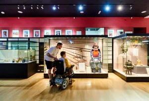 A person looks at a display of Japanese arms and armour with a child who is in a wheelchair.
